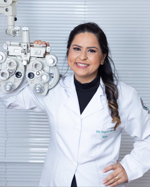 Dra. Bruna Macedo, oftalmologista, de jaleco branco, sorrindo, com o refrator apoiado no ombro